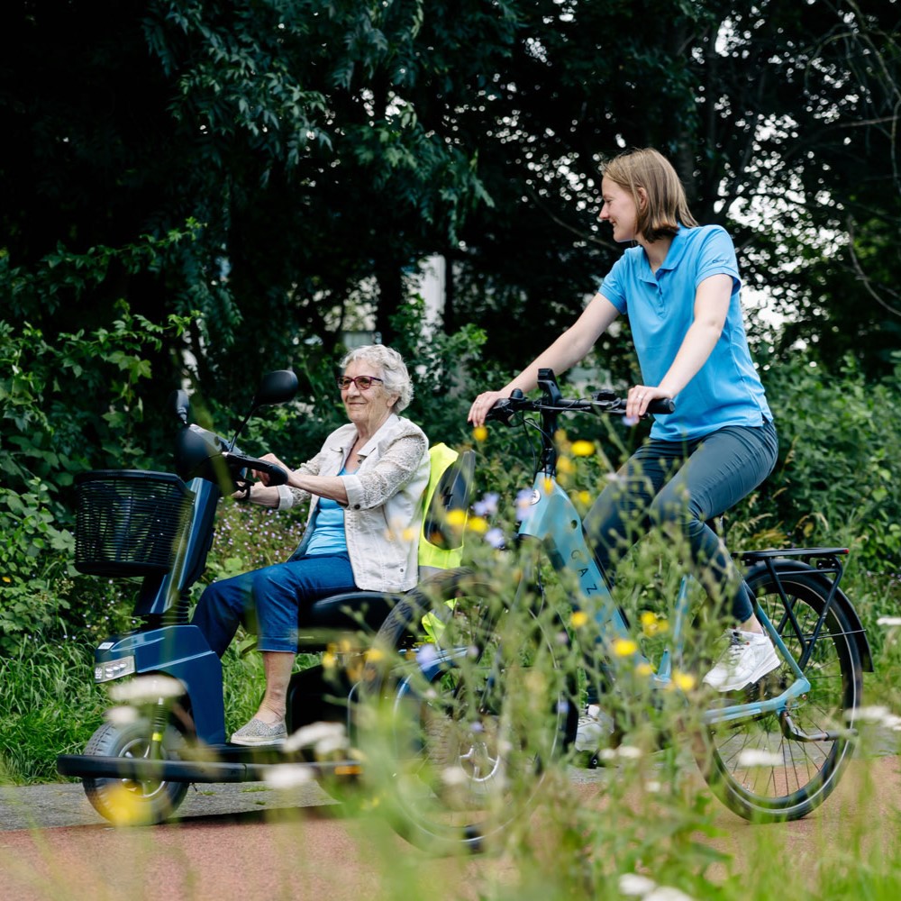 Op de foto is onze ergotherapeut te zien die op de fiets zit, daarnaast haar patiënt in een scootmobiel.