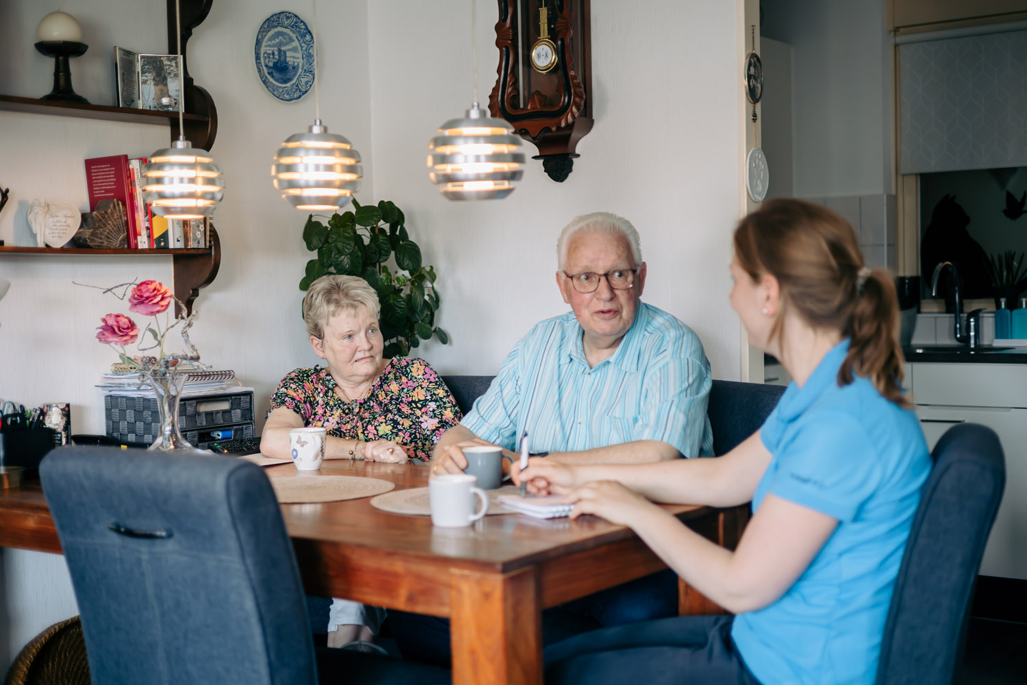 Op de foto is onze ergotherapeut te zien die glimlachend kijkt naar haar patiënt en partner. Ze zitten aan de keukentafel met elkaar.