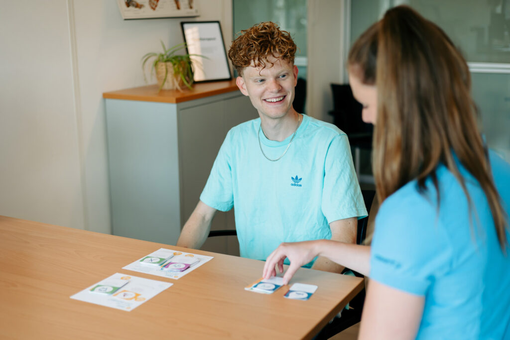 Op de foto staat een jongen, een cliënt, die kijkt naar zijn ergotherapeut. De jongen heeft een glimlach op zijn gezicht en heeft een blauw shirt aan.