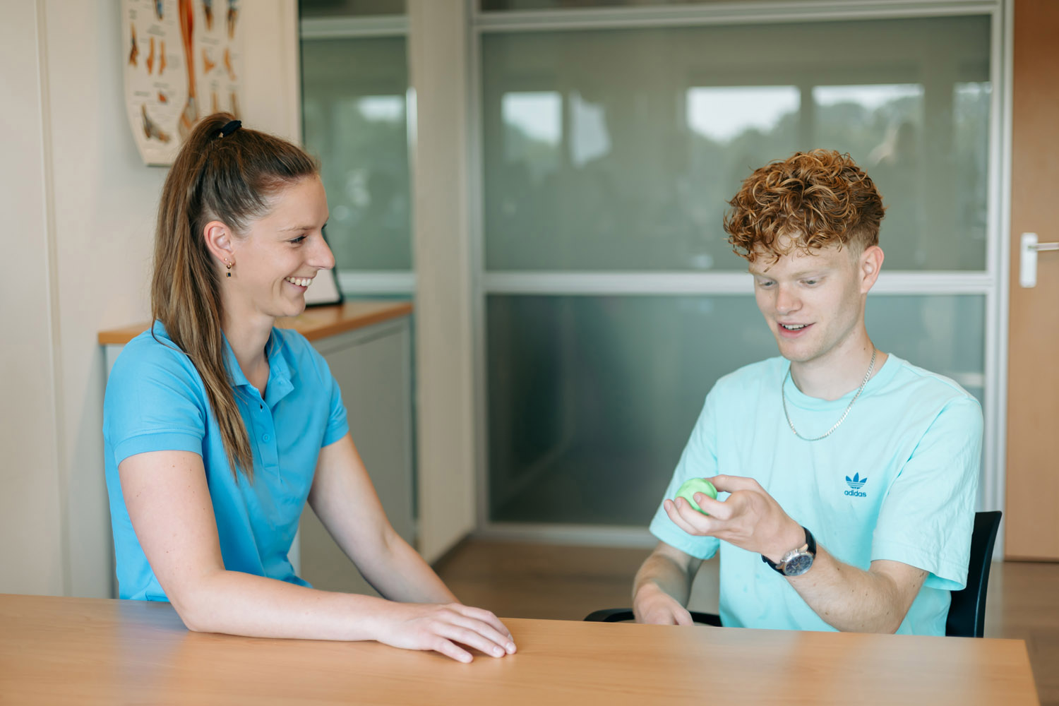 Op de foto is onze ergotherapeut te zien die met een glimlach kijkt naar haar patiënt. Dit is een jongen van ongeveer 18 jaar. Hij heeft een balletje in zijn handen waar hij naar kijkt.