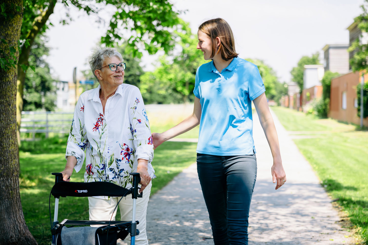 Op de foto is onze ergotherapeut te zien die samen met een cliënt aan het wandelen is buiten. De patiënt loopt met een rollator.