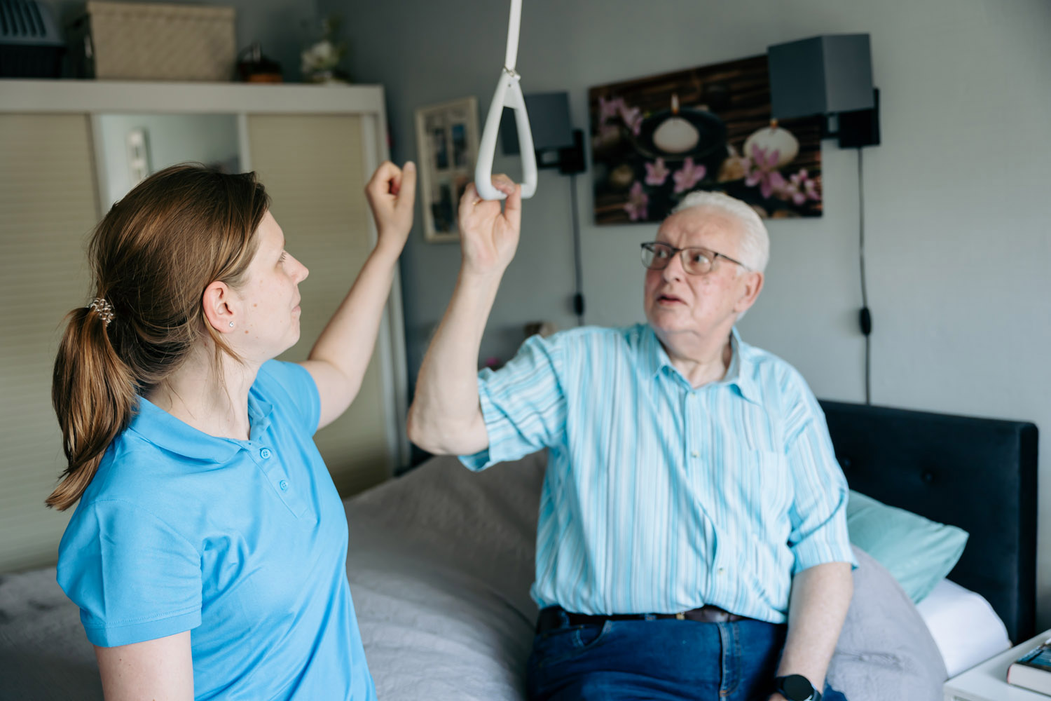 Op de foto staat onze ergotherapeut de slaapkamer van een cliënt. De man, de patiënt zit op bed en houd zich vast een beugel die boven zijn bed hangt om goed te kunnen opstaan vanuit bed.